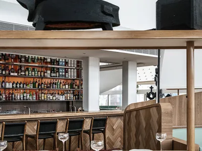 Modern restaurant interior with wooden tables, metal stools, and glass shelves displaying liquor.