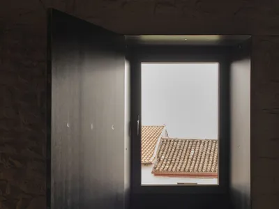 A minimalist bedroom with hardwood flooring, a bed dressed in light gray linens, a book on the windowsill, and a solitary cha