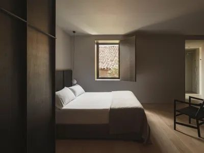 A minimalist bedroom with a bed, chair, and small window overlooking a tiled rooftop.