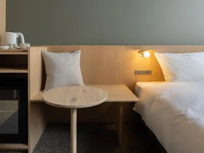 A minimalist bedroom featuring a bed with wooden headboard and nightstand, a small wooden side table, and a wall-mounted lamp