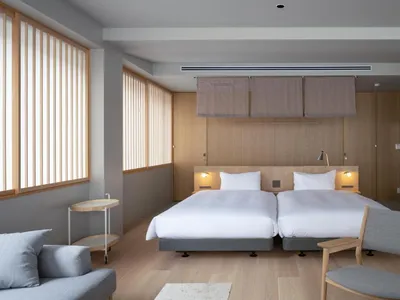 Minimalist hotel room with twin beds, wood furniture, cloth wall coverings, and light fixtures.