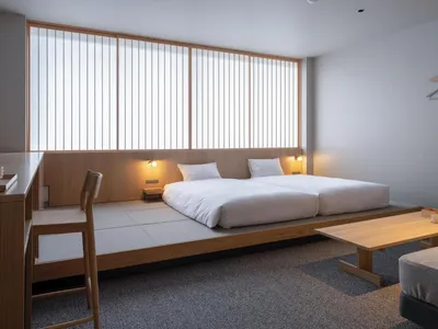 A minimalist bedroom featuring white bedding on a low wooden platform bed, a simple wooden table, and bright natural light en