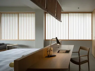 A minimalist bedroom featuring a wooden desk with a lamp, a bed, and a wooden chair.