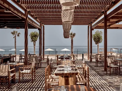 An outdoor restaurant setting with wooden tables, chairs, and a pergola structure overlooking the sea.