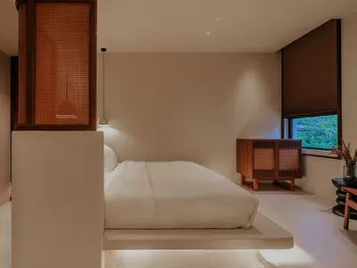 A minimalist bedroom with neutral tones, featuring a bed, side table, and cabinet with a window overlooking greenery.