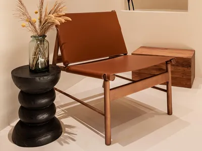 A minimalist indoor lounge area featuring a leather and wooden chair, a vase with dried plants on a black modern table, and a