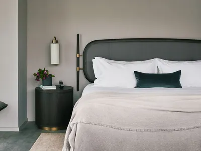 A modern bedroom featuring a fabric bed with a black headboard on a grey floor, accompanied by a side table with a lamp and a
