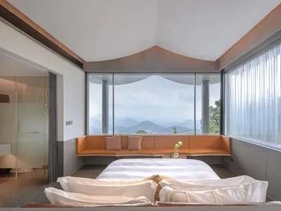 A modern bedroom with a white bed, orange seating area, and large windows overlooking mountains.