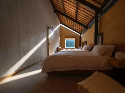 A minimalist bedroom with wooden beams, showing a bed with white bedding and a window with natural light.