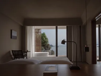 Modern minimalist bedroom with wooden furniture, fabric bedding, and metallic accents overlooking the ocean.
