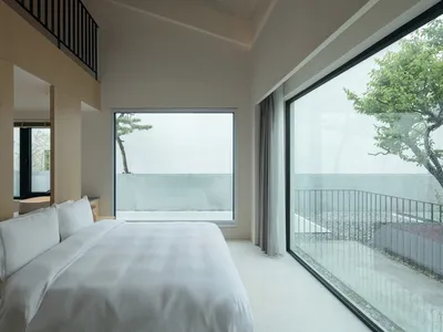 Modern minimalist bedroom featuring a glass window, white bedding, and wooden accents.