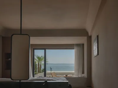 Modern bedroom with concrete walls and floor, large window overlooking the ocean, and minimalist furniture.