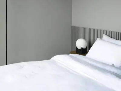 A minimalist bedroom with a white bed, lamp, and grey walls.