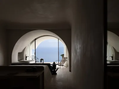 A minimalist bedroom featuring a bed in front of a large arched window overlooking the sea.