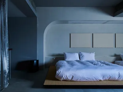 Minimalist bedroom with white bedding on a concrete platform bed, three white square paintings on the wall, and concrete floo