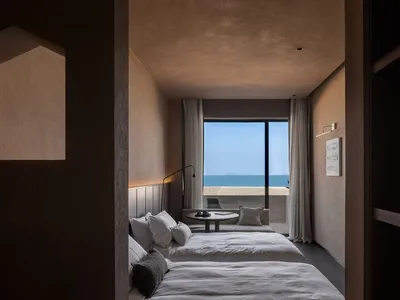A minimalist hotel room with concrete walls and a view of the ocean through floor-to-ceiling windows.
