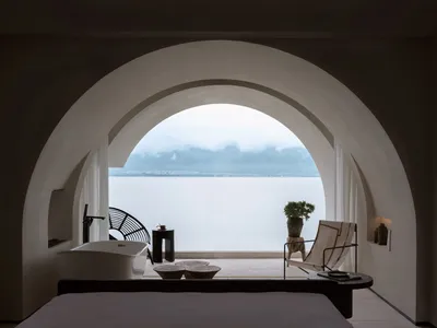 A minimalist hotel suite with a concrete floor, a bathtub, a lounge chair, and a table with a flower pot by the ocean.