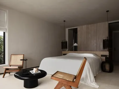 A minimalist bedroom with a large bed dressed in white linens, minimalist wooden furniture, and a black coffee table with a s