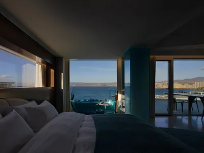 A hotel room interior featuring a glass wall with a view of the ocean and cityscape.