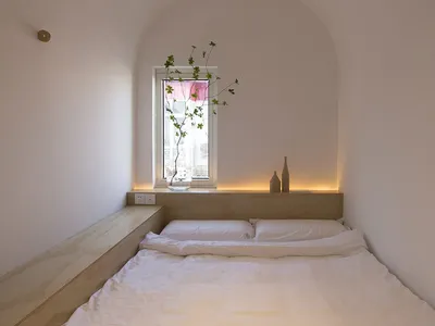 A minimalist bedroom with a white bed and wooden frame, featuring a small window with a plant and two decorative items on the