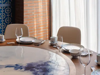 A round wooden table set in an industrial restaurant interior with white curtains and a brick wall.