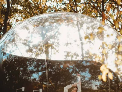 A woman sits on a transparent plastic globe bed outdoors surrounded by trees.