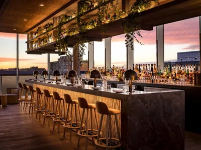 Industrial-style restaurant bar with wooden stools and glass shelves displaying bottles.
