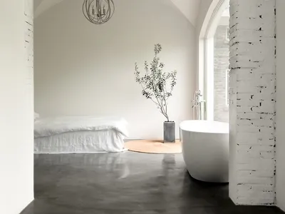 Modern minimalist bedroom with a white bed and a round tree in a vase next to a freestanding bathtub, all under a white brick