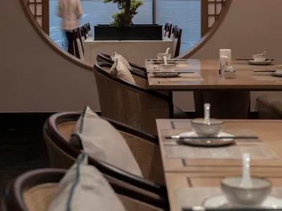 A modern restaurant interior with wooden tables and chairs, circular window, and bonsai tree.