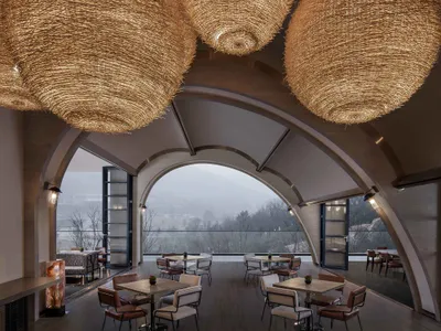 Modern restaurant lounge area with wooden furniture and glass panels, featuring a large window offering scenic views.