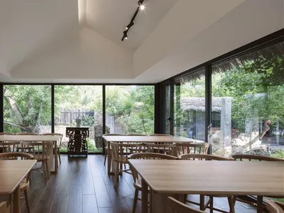 A spacious modern restaurant interior features large windows with a view of greenery, wooden tables, and glass railings.
