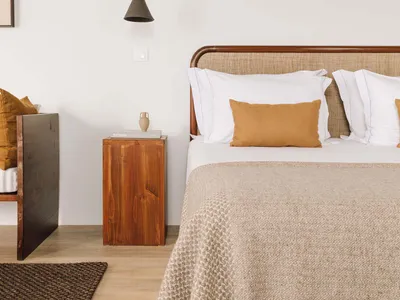 A minimalist bedroom featuring a white bed with a beige textured blanket, white pillows, and a wooden nightstand with a brown