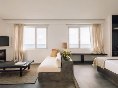 Modern hotel room featuring a concrete sofa, fabric cushions, and wooden elements, with a view of the ocean through large win