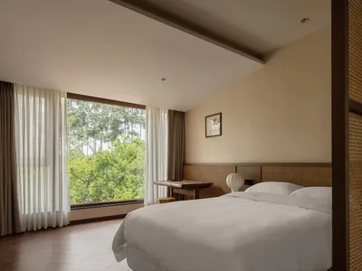 A minimalist bedroom featuring a double bed with a white textured headboard, white bedding, and large windows with sheer curt