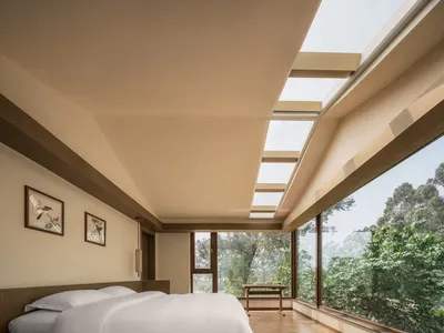 Minimalist bedroom with a white bed, wooden flooring, and large windows overlooking greenery.