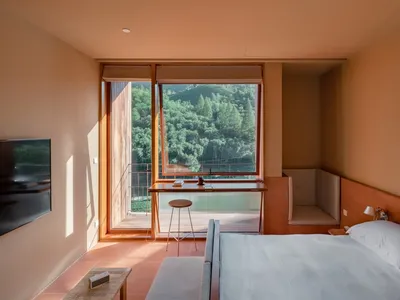 A minimalist bedroom with a large window overlooking a forest, featuring a bed with a wooden headboard and white bedding, and