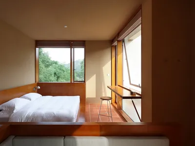 A modern minimalist bedroom featuring a wooden bed with white bedding, natural light, and a view of greenery through large wi