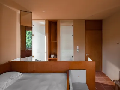 A modern minimalist bedroom featuring a bed, two chairs, and a television in a minimalist design with wooden and textile elem