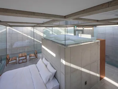 A minimalist bedroom featuring a platform bed with a glass railing, surrounded by concrete walls and floors.