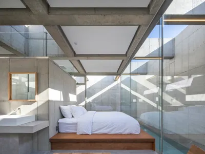 Modern minimalist bedroom with concrete walls, a raised bed platform, and a glass wall showcasing a pool.