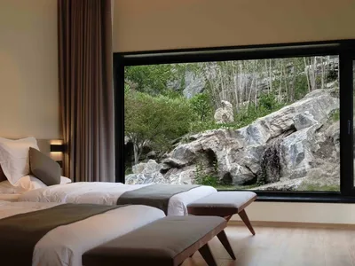 A well-lit hotel room with a view of greenery and rocks outside through a large window.