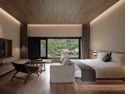 Modern minimalist bedroom featuring a large bed with white sheets, a white sofa, and a wooden floor and ceiling. The room has