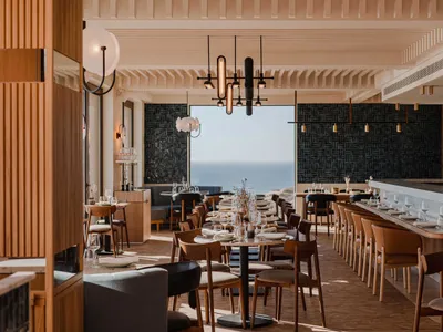 Modern restaurant interior with sea view, featuring wooden tables, seating, and minimalist decor.