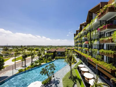 A modern hotel with concrete and glass exteriors, featuring a large pool area and lush greenery.