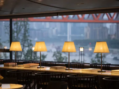A well-lit cafe interior with wooden tables and chairs, warm lighting, and a view of a bridge over a body of water.