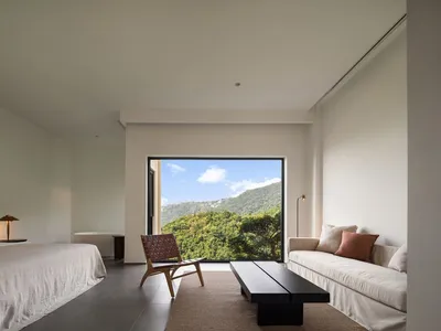 A minimalist bedroom with a bed, sofa, and large window showcasing a scenic view of mountains and trees.