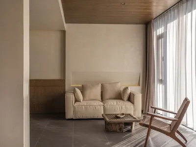 A minimalist living room with a brown sofa, beige cushions, and a wooden coffee table in front of a window with white curtain