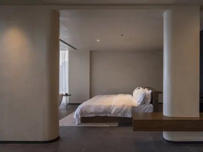 A minimalist bedroom with concrete walls and floor, featuring a bed with white bedding and wooden accents.