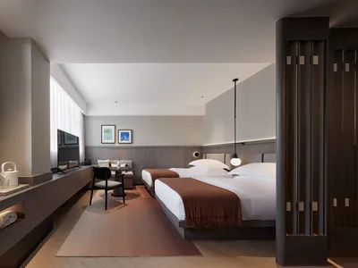 A minimalist bedroom with a bed covered in a brown throw blanket, a small table with a vase, and a chair in front of it.