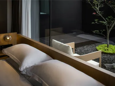 Modern minimalist bedroom with a wooden headboard and white bedding, featuring a small indoor garden with a tree and rocks.